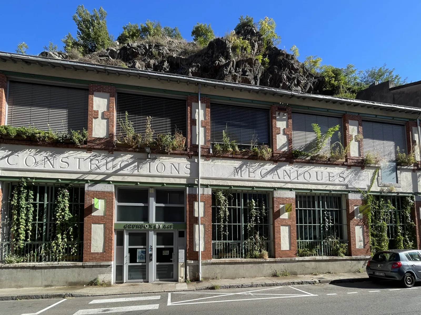 Façade de la grünboX à Vienne : ancienne usine en brique et enduit, jardinières aux fenêtres, inscription « Constructions mécaniques », rocher boisé en arrière-plan