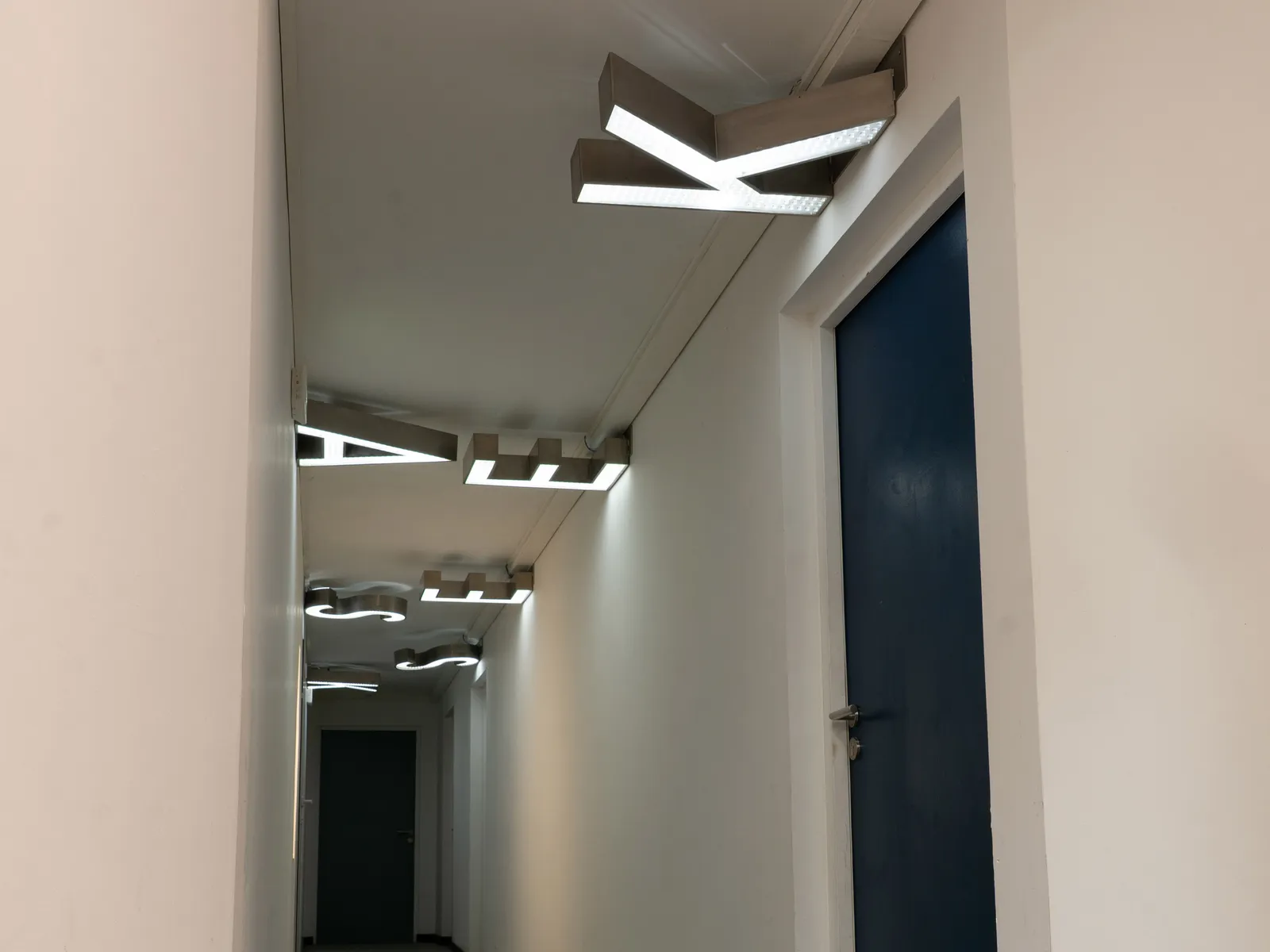 Couloir de la grünboX après travaux : murs blancs, porte bleue et luminaires suspendus au plafond